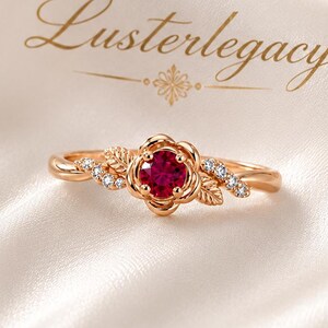 May include: A rose gold ring featuring a central round-cut red gemstone surrounded by metal petals and small clear stones on the band. The text Lusterlegacy is printed above the ring. The design includes leaf-like details on the sides of the setting.