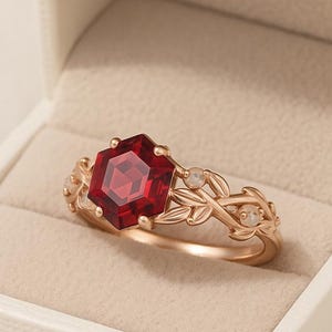 Könnte beinhalten: Ein roségoldener Ring mit einem sechseckigen, tiefroten Edelstein. Der Ring hat ein florales Design mit kleinen, runden Akzentsteinen. Der Ring wird in einer weißen Schmuckschatulle präsentiert.