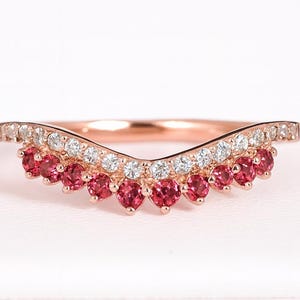 Könnte beinhalten: Ein roségoldener Ring mit geschwungenem Design. Der Ring hat eine Reihe kleiner, runder, roter Edelsteine am unteren Rand und eine Reihe kleiner, klarer Edelsteine am oberen Rand. Der Ring ist vor einem weißen Hintergrund platziert.
