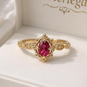 Pode incluir: Um anel de ouro com uma pedra preciosa rosa-escura de corte oval, engastada numa armação ornamentada de estilo filigrana. A banda inclui pequenas pedras redondas e claras. O anel é apresentado numa caixa branca com o texto Lusterlegacy impresso no interior.