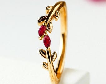 May include: A gold-colored ring featuring a leaf design with two red gemstones. The ring is set against a white background, highlighting its intricate details and vibrant color.