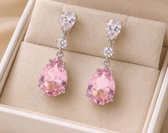 May include: A pair of silver-toned drop earrings featuring a small round clear stone above a large pear-shaped pink stone, presented in a white box with the gold text Lusterlegacy printed on the inside lid, accompanied by small white flowers.