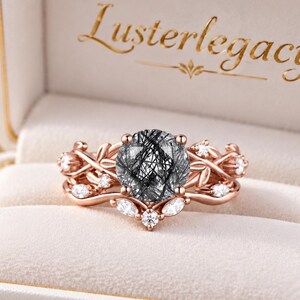 May include: A rose gold ring featuring a round rutilated quartz centre stone with black needle-like inclusions, set in a band designed with metal leaves and small white accent stones, presented in a cream box with the text Lusterlegacy.