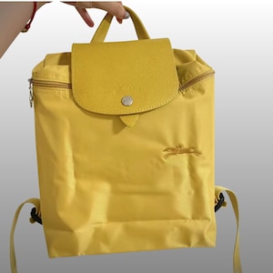 May include: A bright yellow backpack featuring a textured leather flap with a silver-toned snap button closure. The bag has a top handle, adjustable shoulder straps, and an embroidered horse logo on the front. The main compartment is secured with a zipper.