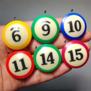 Op de afbeelding: Zes ronde knopen of cabochons in biljartbalstijl met de nummers 6, 9, 10, 11, 14 en 15. Elk stuk heeft een crèmekleurig midden met een zwart cijfer en een gekleurde rand in respectievelijk geel, groen, blauw, rood, groen en roze.