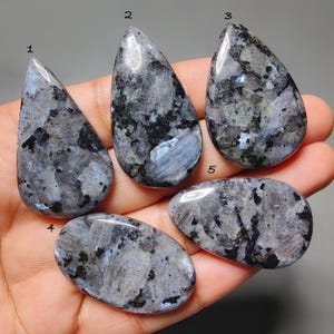 May include: Five polished labradorite gemstones in various shapes, including teardrop and oval. The stones display a mix of grey, black, and blue hues with a shimmering effect. The gemstones are held in a hand.