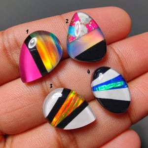 May include: Four teardrop-shaped gemstones with unique designs. One features pink, black, and iridescent colors. Another has pink, blue, and gold accents. The third has white, black, and orange stripes. The last has black, white, and blue stripes.