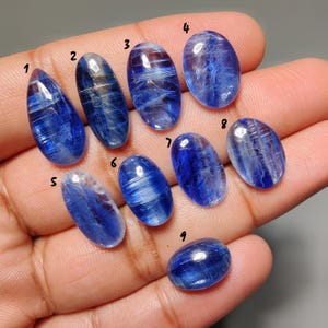 May include: A collection of nine polished blue kyanite gemstones in various oval and teardrop shapes. The stones display a range of blue hues with visible internal patterns. The gemstones are held in a hand, showcasing their natural beauty and variations.