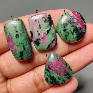 Op de afbeelding: Vier gepolijste robijn in zoisiet edelstenen in verschillende vormen. De stenen hebben een mix van groene, roze en zwarte kleuren. De vormen omvatten een rechthoek, ovaal en druppel. De edelstenen worden in een hand gehouden.