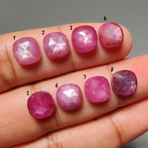 May include: Eight faceted, cushion-cut rubies in varying shades of pink and red. The gemstones are displayed on a hand, showcasing their polished surfaces and internal structures. Each stone is numbered for identification, highlighting their unique characteristics.