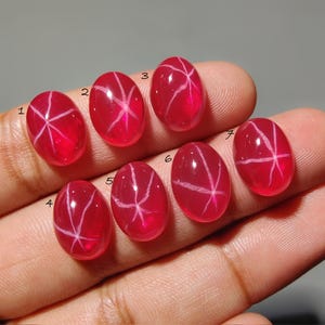 May include: Seven oval-shaped, polished red gemstones with a star-like pattern of white lines. The gems are displayed on a hand, showcasing their vibrant color and unique internal designs. These gemstones are likely used for jewelry.