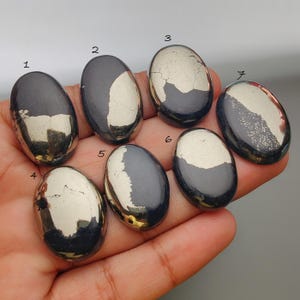 May include: Seven polished pyrite stones, each with a unique pattern of metallic gold and dark grey. The stones are oval-shaped and vary in the amount of gold veining. The stones are held in a hand against a neutral background.