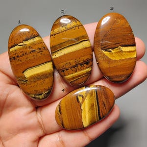May include: Four polished oval tiger's eye gemstones. The stones display a mix of brown, gold, and black bands. Each stone has a unique pattern and shape, with a smooth, glossy finish. The gemstones are held in a hand.