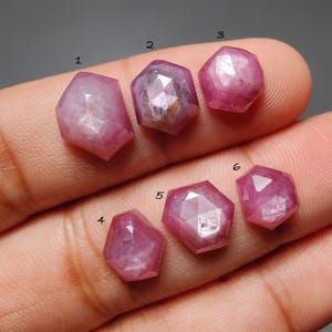 以下が含まれることがあります： さまざまな形とサイズの6つのファセット加工されたピンク色の宝石が表示されています。宝石は半透明で、内部構造とインクルージョンが見えます。石は手で持たれており、色と透明度を示しています。