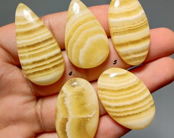 Pode incluir: Cinco cabochões de ágata com faixas amarelas e brancas rotulados com os números de um a cinco. As pedras apresentam superfícies lisas e arredondadas com camadas minerais horizontais e verticais distintas em tons de creme, ouro e branco translúcido.