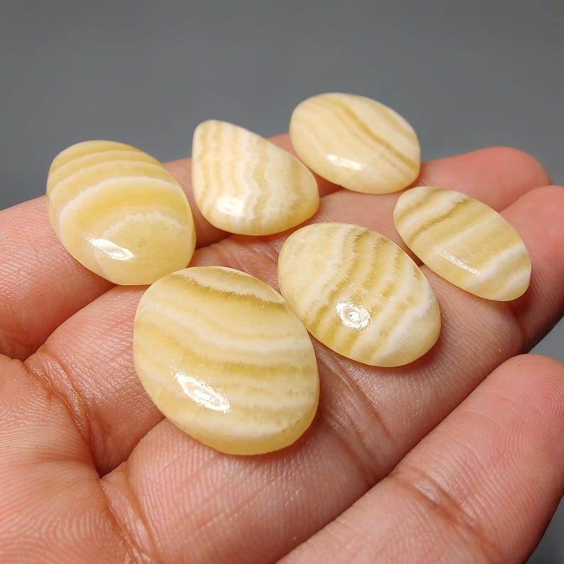May include: A collection of seven polished, oval-shaped gemstones. The stones display a creamy yellow color with distinctive, parallel, light-colored banding patterns. The gemstones are smooth and appear to be held in a hand.