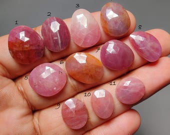 Puede incluir: Una mano muestra once piedras preciosas facetadas en tonos rosa, rojo y naranja. Las gemas tienen varias formas, incluyendo cortes ovalados y redondos. Las piedras se muestran sobre un fondo neutro.