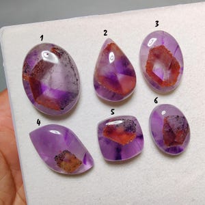 Pode incluir: Seis pedras preciosas de ametista polidas de várias formas. As pedras exibem uma mistura de tons roxos e castanho-avermelhados. Cada pedra tem um padrão único sobre um fundo branco. As pedras preciosas são provavelmente para fazer joias ou colecionar.