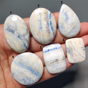 May include: A collection of six polished gemstones in various shapes: oval, teardrop, round, and rectangular. The stones feature a mix of white, blue, and tan colors, with unique veining patterns. The gemstones are held in a hand.