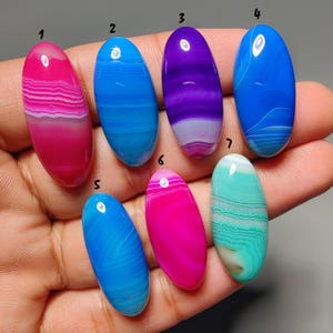 Op de afbeelding: Zeven ovale edelstenen in verschillende kleuren, waaronder roze, blauw, paars en turquoise. Elke gepolijste steen vertoont unieke bandpatronen. De edelstenen worden in een hand gehouden, om hun grootte en vorm te laten zien.