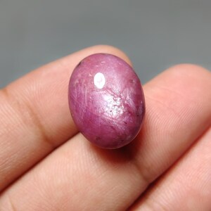 May include: An oval-shaped, cabochon-cut gemstone with a deep magenta and pink hue. The surface of the stone displays a visible star-like optical phenomenon known as asterism, characterized by intersecting light rays across the polished surface.
