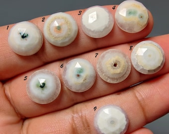 Puede incluir: Nueve piedras preciosas redondas y facetadas dispuestas en una cuadrícula, numeradas del uno al nueve. Estas piedras presentan una base blanca opaca con variadas inclusiones centrales en tonos verde, naranja y bronce, con un patrón circular.