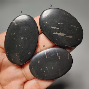 Pode incluir: Três pedras ovais polidas, de cor cinza escuro. Apresentam inclusões claras sutis e uma superfície lisa. Estas pedras parecem ser pedras preciosas naturais, possivelmente usadas para joias ou decoração. Cada pedra mede cerca de 5-7 cm.