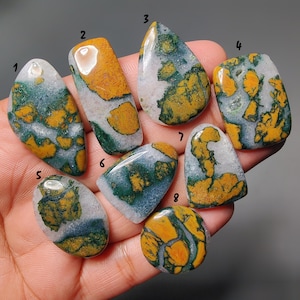 May include: A collection of eight polished gemstones with unique patterns. The stones feature a mix of colors, including green, yellow, and white, with various shapes and sizes. The gemstones are held in a hand, showcasing their natural beauty and intricate designs.