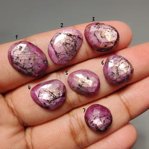 Può includere: Sette gemme di rubino lucide, di forma ovale e di varie dimensioni, sono mostrate su una mano. I rubini variano di colore dal rosa intenso al viola rossastro, con inclusioni più scure. Ogni pietra ha una forma e una superficie uniche.