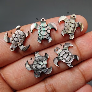 Puede incluir: Cinco pequeños colgantes iridiscentes con forma de tortuga. Cada colgante tiene un patrón de caparazón detallado y un lazo para sujetarlo a la joyería. Los colgantes son plateados y tienen un aspecto nacarado.