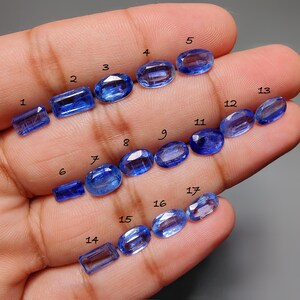 May include: A collection of seventeen blue kyanite gemstones of varying shapes and sizes, including oval, rectangular, and rounded forms. The gemstones are displayed on a hand, showcasing their clarity and color.