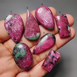 Pode incluir: Uma coleção de sete pedras preciosas de rubi polidas em várias formas, incluindo gota, oval e retangular. As pedras exibem um tom rosa-avermelhado vibrante com inclusões pretas e verdes. As pedras preciosas são seguradas em uma mão.
