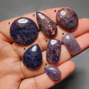Peut inclure: Sept pierres précieuses polies en cabochon dans des tons de violet profond avec des inclusions métalliques. Les pierres sont numérotées de un à sept et varient en forme, incluant des formes rondes, en goutte, rectangulaires et ovales lisses.