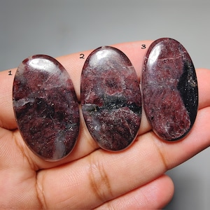May include: Three oval-shaped polished gemstones featuring deep burgundy and dark red hues with black and translucent white inclusions. Each stone is marked with a small black number, labeled 1, 2, and 3, indicating distinct individual pieces.