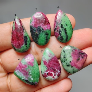 Puede incluir: Seis piedras preciosas pulidas de rubí en zoisita de varias formas. Las piedras presentan una mezcla de colores rojo, verde y negro. Las piedras preciosas se sostienen en una mano, mostrando su belleza natural y patrones únicos.