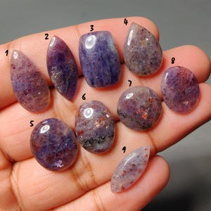 Peut inclure: Une collection de neuf pierres précieuses polies de formes variées, dont des gouttes, des ovales et des rectangles. Les pierres présentent un mélange de teintes violettes et rouges, avec une qualité translucide. Les pierres sont tenues dans une main, mettant en valeur leur beauté naturelle.