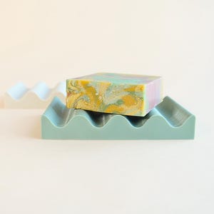 May include: A colorful bar of soap rests on a teal soap dish with a wavy design. A second white soap dish is in the background. The soap has yellow, green, and purple hues.