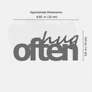 K&ouml;nnte beinhalten: Graues Holzschild mit den ausgeschnittenen Worten "hug often". Das Schild ist ca. 22 cm breit und 10 cm hoch. Der Hintergrund ist wei&szlig;.