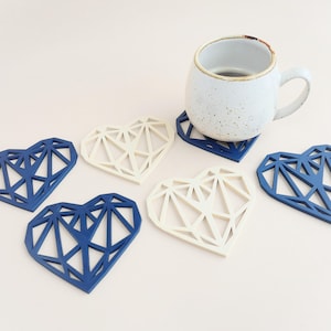 May include: A white ceramic mug rests on a geometric heart-shaped coaster. Several other heart-shaped coasters in navy blue and off-white are scattered around. The coasters have a modern, angular design, suitable for home decor.
