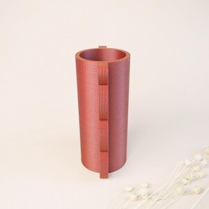 May include: A cylindrical, reddish-brown vase with a textured surface. A vertical, rectangular design is attached to the side of the vase. The vase is empty and stands on a light-colored surface. Decorative dried flowers are in the background.