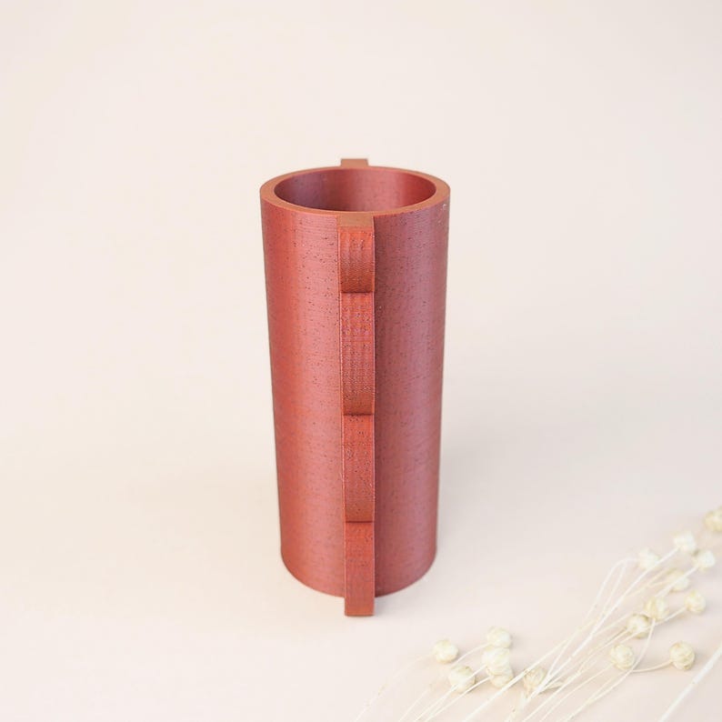 May include: A cylindrical, reddish-brown vase with a textured surface. A vertical, rectangular design is attached to the side of the vase. The vase is empty and stands on a light-colored surface. Decorative dried flowers are in the background.