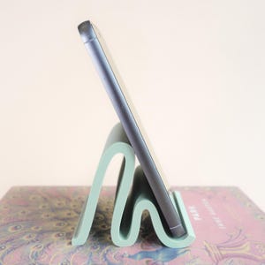 May include: A mint green phone stand with a wavy design, holding a gray smartphone. The stand is made of a solid material and sits on a book with a peacock feather pattern.