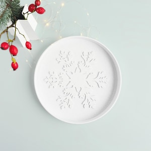May include: A white, round decorative tray featuring a snowflake design. The tray is made of a textured material and is set against a light blue background. Red berries and a sprig of greenery are in the upper left corner, with string lights.