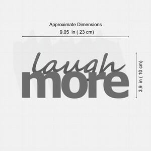 K&ouml;nnte beinhalten: Graue Holz-Wanddekoration mit den Worten "laugh more". Das Wort "laugh" ist in einer Schreibschrift &uuml;ber dem Wort "more" in Blockschrift geschrieben. Die Ma&szlig;e betragen ca. 23 cm Breite und 10 cm H&ouml;he.