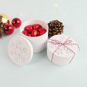 May include: White, round gift boxes with snowflake cutouts and red and white twine. One box is open, revealing red heart-shaped candies. Christmas ornaments and a pinecone are also in the image.