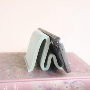 May include: A light green, wavy phone stand holding a silver smartphone. The stand has a unique, curved design and is made of a solid material. The phone is positioned upright, resting against the stand. The stand is on a book with a floral pattern.