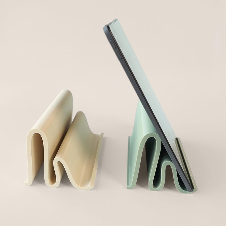 May include: Two wavy, minimalist tablet stands in cream and mint green. One stand holds a tablet. The stands are made of a smooth, solid material and have a unique, folded design. The background is a soft, neutral color.