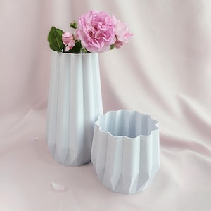 Può includere: Due vasi azzurri, con finitura strutturata. Uno è alto e sottile, con rose rosa. L'altro è un vaso più corto e rotondo. Entrambi hanno un design a coste e geometrico. Lo sfondo è un tessuto rosa tenue.
