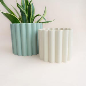 Puede incluir: Dos macetas decorativas con un diseño ondulado de costillas verticales. Una es de un verde suave y apagado, y la otra es de un blanco roto cremoso. Las macetas están vacías, listas para plantas. El fondo es rosa claro.