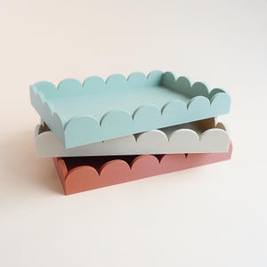 May include: Three rectangular wooden trays stacked on top of each other. The top tray is light blue, the middle tray is white, and the bottom tray is a muted coral color. Each tray has a scalloped edge.